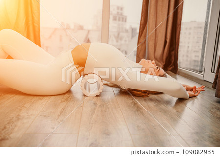Adult athletic woman, in white bodysuit, performing fascia exercises on the floor - caucasian woman using a massage foam roller - a tool to relieve tension in the back and relieve muscle pain - the 109082935