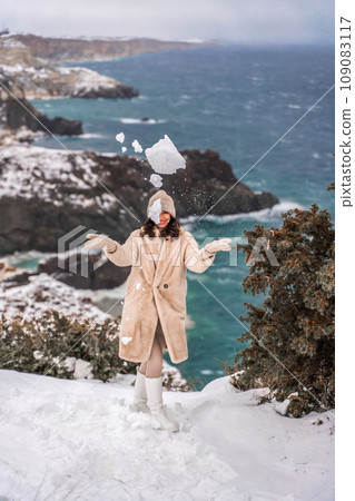 Woman snow sea. Amidst a wintry backdrop, a woman in a beige faux fur coat gleefully engages in a snowball fight, adding joy to the serene seascape. 109083117