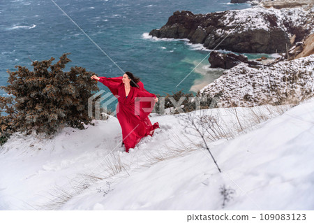 Woman red dress snow sea. Happy woman in a red dress in the snowy mountains by the emerald sea. The wind blows her clothes, posing against sea and snow background. Woman red dress snow sea. Happy woman in a red dress in the snowy mountains by the emerald sea. The wind blows her clothes, posing against sea and snow background. 109083123