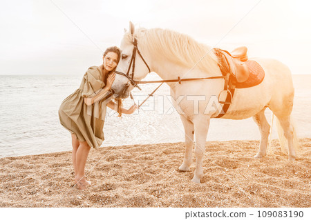 A woman in a dress stands next to a white horse on a beach, with the blue sky and sea in the background. A woman in a dress stands next to a white horse on a beach, with the blue sky and sea in the background. 109083190
