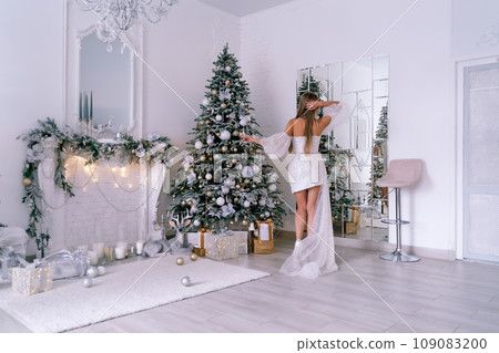 The blonde in the Christmas room. A beautiful blonde woman in a shiny light short dress with a train stands in a beautiful bright room decorated with a festive interior with a Christmas tree. 109083200