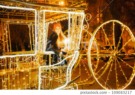 Woman holding sparkler night while celebrating Christmas outside. Dressed in a fur coat and a red headband. Blurred christmas decorations in the background. Selective focus Woman holding sparkler night while celebrating Christmas outside. Dressed in a fur coat and a red headband. Blurred christmas decorations in the background. Selective focus 109083217