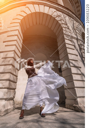 Stylish woman building city. A dancer in a long white skirt dances in front of a building with an arch. The skirt develops in the wind. 109083220