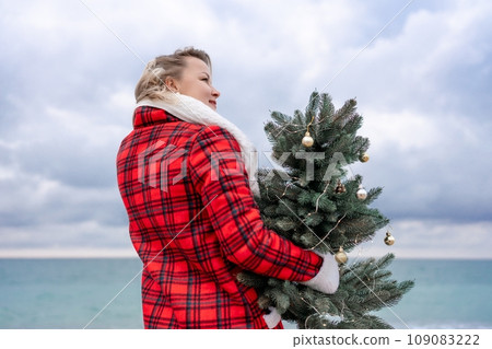 Blond woman holding Christmas tree by the sea. Christmas portrait of a happy woman walking along the beach and holding a Christmas tree in her hands. Dressed in a red coat, white suit. Blond woman holding Christmas tree by the sea. Christmas portrait of a happy woman walking along the beach and holding a Christmas tree in her hands. Dressed in a red coat, white suit. 109083222