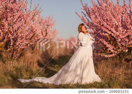 Woman blooming peach orchard. Against the backdrop of a picturesque peach orchard, a woman in a long white dress enjoys a peaceful walk in the park, surrounded by the beauty of nature. 109083231