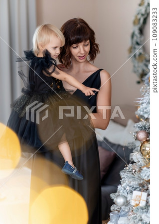 Mother daughter 2 years old Christmas tree. Both are dressed in black dresses, the mother holds the girl in her arms and both look at each other. The family celebrates Christmas. Mother daughter 2 years old Christmas tree. Both are dressed in black dresses, the mother holds the girl in her arms and both look at each other. The family celebrates Christmas. 109083233