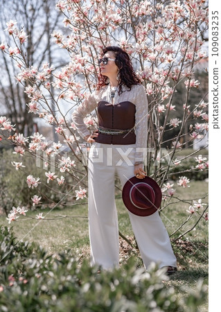 Magnolia park woman. Stylish woman in a hat stands near the magnolia bush in the park. Dressed in white corset pants and posing for the camera. Magnolia park woman. Stylish woman in a hat stands near the magnolia bush in the park. Dressed in white corset pants and posing for the camera. 109083235