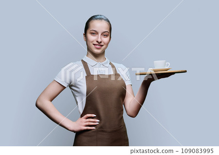 Young female waitress in apron with tray of cup of coffee on gray background 109083995