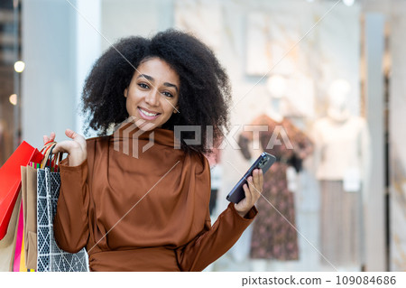 Portrait of a young beautiful woman in a supermarket, a large clothing store, smiling, looking at the camera, holding a phone and colorful packages with shopping gifts. 109084686