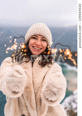 Outdoor winter portrait of happy smiling woman, light faux fur coat holding heart sparkler, posing against sea and snow background Outdoor winter portrait of happy smiling woman, light faux fur coat holding heart sparkler, posing against sea and snow background 109084688