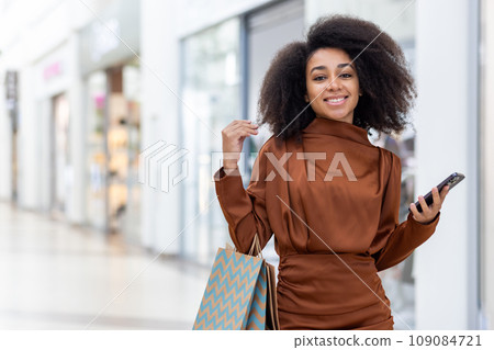 Portrait of a young beautiful woman in a supermarket, a large clothing store, smiling, looking at the camera, holding a phone and colorful packages with shopping gifts. 109084721