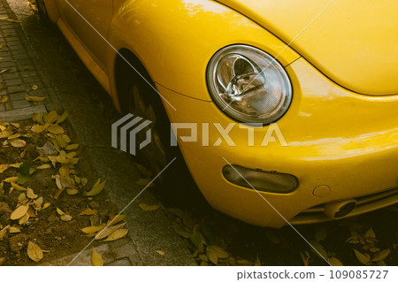 Madrid, Spain November 22, 2023 Volkswagen New Beetle. Compact bright yellow car. Top view of a car's bumper and headlights on the autumnal city street. Fall season and fallen leaves on a ground. 109085727