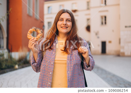 Attractive young female 30s tourist with backpack is holding prezel, traditional polish snack on the Market square in Krakow. Traveling Europe in spring. Selective focus. High quality photo 109086254