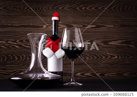 Decanter, glass and bottle wearing Santa Claus hat and scarf with red wine on wooden rustic background. Christmas celebration. New year decoration Decanter, glass and bottle wearing Santa Claus hat and scarf with red wine on wooden rustic background. Christmas celebration. New year decoration 109088125