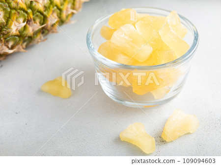 Dried soft sweet pineapple slices in glass bowl with raw pineapple on light table.Macro 109094603