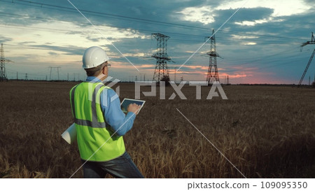 Manager in helmet types on tablet examining power transmission lines in wheat field. Electrician with drawings and helmet controls power distributions substation equipment via tablet in sunset field Manager in helmet types on tablet examining power transmission lines in wheat field. Electrician with drawings and helmet controls power distributions substation equipment via tablet in sunset field 109095350