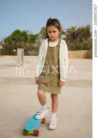 Upset Caucasian child girl standing on her skateboard on one leg on skatepark, expressing sad emotions looking at camera Upset Caucasian child girl standing on her skateboard on one leg on skatepark, expressing sad emotions looking at camera 109097267