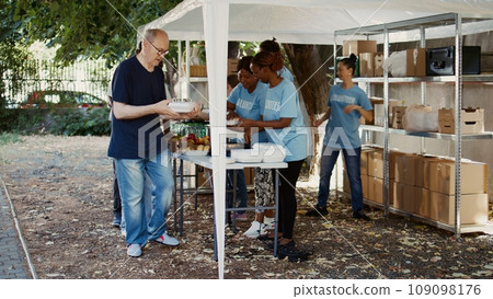 Scene showcasing group of volunteers giving food donations to less privileged. Young voluntary individuals volunteering their time to help the needy and provide homeless people free meals. Handheld. 109098176