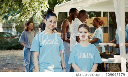 Portrait shot of mother and daughter participating in hunger relief program at outdoor food bank. Caucasian volunteers in blue t-shirts and looking at camera, ready to assist the needy. Tripod shot. 109098179