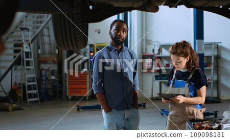 Meticulous technician in garage workplace stands with customer underneath suspended car, looking together for replacement parts. Expert helps client mend automobile using tablet to find components 109098181