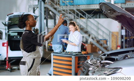 Licensed engineer in auto repair shop using augmented reality hologram to visualize car components in order to fix them. African american woman using AR technology while working on damaged vehicle 109098200