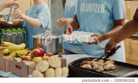 Multiethnic people in blue t-shirts, assembled outside to hand out food donations and help the homeless and hungry. Focus on volunteers serving free, freshly prepared meals to needy individuals. Multiethnic people in blue t-shirts, assembled outside to hand out food donations and help the homeless and hungry. Focus on volunteers serving free, freshly prepared meals to needy individuals. 109098208