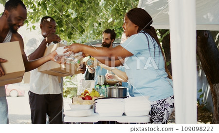 In a sunny day, multiethnic volunteers distribute donations of food to the needy, offering help and hope to homeless people. Caring charity workers giving hunger relief packages to the less fortunate. 109098222