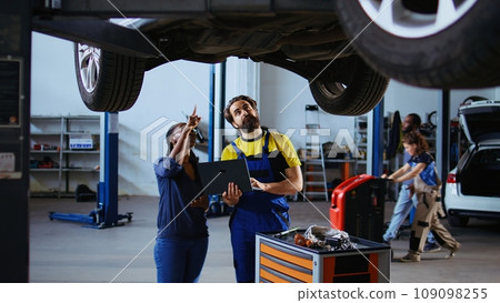 Serviceman and customer standing underneath car in garage using laptop to order new parts after finding damages. Employee assisting client by looking online for replacing automobile components 109098255
