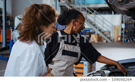 Trained repairman in garage workspace fixing car for customer, thoroughly checking underneath vehicle hood. African american expert doing annual maintenance on client automobile in auto repair shop Trained repairman in garage workspace fixing car for customer, thoroughly checking underneath vehicle hood. African american expert doing annual maintenance on client automobile in auto repair shop 109098264