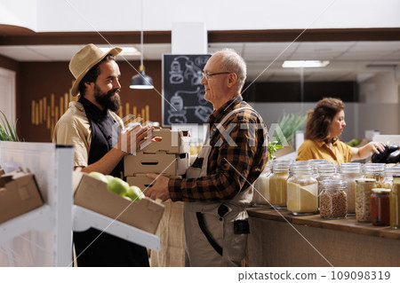 Farmer providing locally grown ecofriendly vegetables to seller selling it to environmentally conscious customers in zero waste shop. Older man handing crates full of vegan produce to retailer 109098319