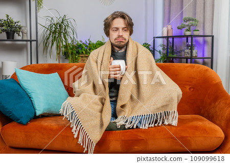 Sick ill Caucasian man wrapped in plaid sit shivering from cold drinking hot coffee or tea at home 109099618