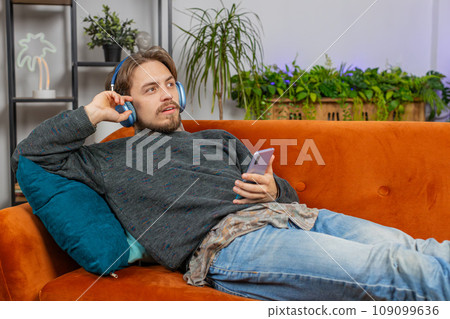 Happy man in wireless headphones relaxing listening favorite rock n roll music lying on home couch 109099636