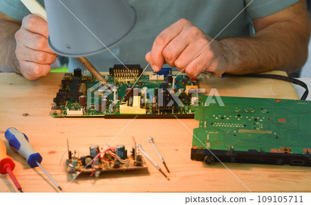 A man holds a soldering iron in his hand and solders a microcircuit A man holds a soldering iron in his hand and solders a microcircuit 109105711