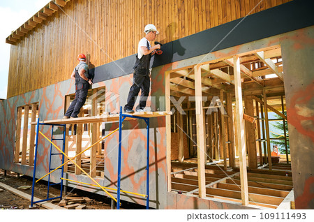 Carpenters constructing wooden framed house. Two men workers cladding facade of house with cement particle boards, fastening them with screwdrivers. Concept of modern eco-friendly construction. Carpenters constructing wooden framed house. Two men workers cladding facade of house with cement particle boards, fastening them with screwdrivers. Concept of modern eco-friendly construction. 109111493