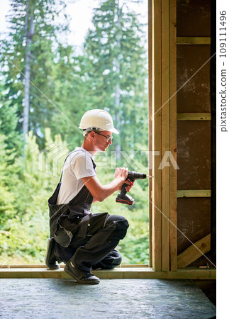 Carpenter constructing wooden framed house. Man worker in glasses working with screwdriver, wearing work overalls and helmet. Concept of modern eco-friendly construction. Carpenter constructing wooden framed house. Man worker in glasses working with screwdriver, wearing work overalls and helmet. Concept of modern eco-friendly construction. 109111496
