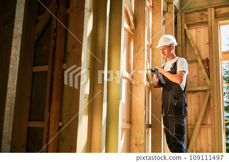Carpenter constructing wooden framed house. Man worker working with screwdriver, wearing work overalls and helmet. Concept of modern eco-friendly construction. Carpenter constructing wooden framed house. Man worker working with screwdriver, wearing work overalls and helmet. Concept of modern eco-friendly construction. 109111497