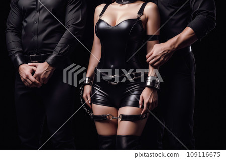 A man standing near imprisoned woman in handcuffs and chains, wearing a sexy black leather bdsm costume isolated on black 109116675