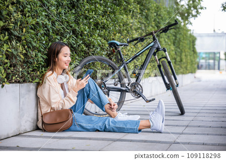 On splendid spring day in park cute young woman is sitting outdoors with her bicycle using her mobile phone to listen to music with earphones. Her joyful typing and bright smile reflect pure happiness On splendid spring day in park cute young woman is sitting outdoors with her bicycle using her mobile phone to listen to music with earphones. Her joyful typing and bright smile reflect pure happiness 109118298