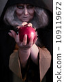Stepmother witch gives poisoned red apple.Woman as witch in black offers red apple as symbol of temptation, poison. Fairy tale, white snow wizard concept. Spooky Halloween, cosplay. 109119672