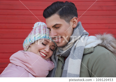 Happy couple in warm clothes near red wooden wall outdoors. Christmas season 109120453