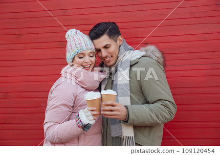 Happy couple in warm clothes with drinks near red wooden wall outdoors. Christmas season Happy couple in warm clothes with drinks near red wooden wall outdoors. Christmas season 109120454