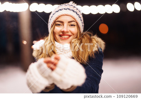 Cheerful young woman holding sparkler in hand in winter forest. Happy cute girl posing with sparkler 109120669