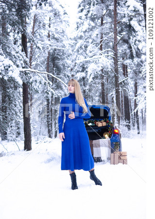 Woman in winter snowy forest in blue dress next to blue car decorated with Christmas decor. Christmas and winter holidays concept. 109121002