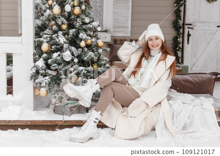 A woman in white winter clothes on the veranda of a country house decorated for the Christmas holidays. Wooden house. 109121017