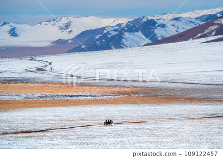 Beautiful Mongolian winter landscape snow mountain with a small figure of a riders on a black horses galloping through a wide snow-covered valley against the backdrop of high mountains. Beautiful Mongolian winter landscape snow mountain with a small figure of a riders on a black horses galloping through a wide snow-covered valley against the backdrop of high mountains. 109122457