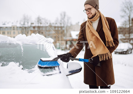 Stylish man in a coat and scarf clears snow from his car during winter snowfall. Seasonality concept Stylish man in a coat and scarf clears snow from his car during winter snowfall. Seasonality concept 109122664