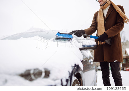 Stylish man in a coat and scarf clears snow from his car during winter snowfall. Seasonality concept Stylish man in a coat and scarf clears snow from his car during winter snowfall. Seasonality concept 109122665