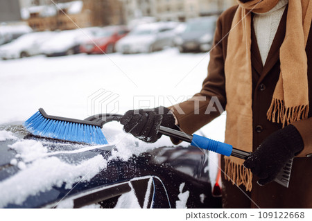 Stylish man in a coat and scarf clears snow from his car during winter snowfall. Seasonality concept 109122668
