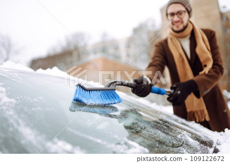 Stylish man in a coat and scarf clears snow from his car during winter snowfall. Seasonality concept Stylish man in a coat and scarf clears snow from his car during winter snowfall. Seasonality concept 109122672