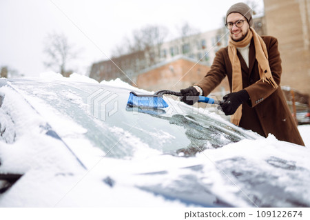 Stylish man in a coat and scarf clears snow from his car during winter snowfall. Seasonality concept Stylish man in a coat and scarf clears snow from his car during winter snowfall. Seasonality concept 109122674